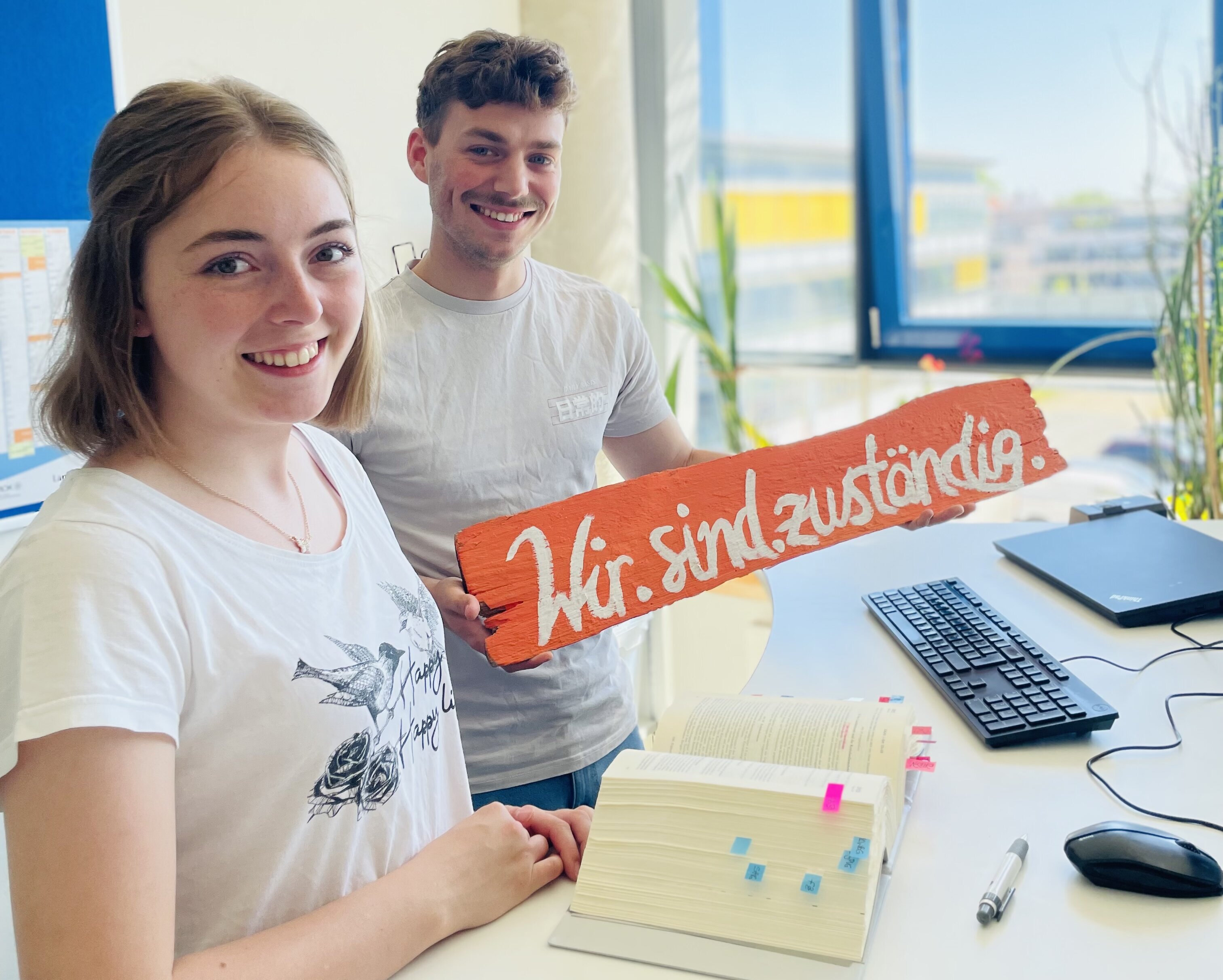Zwei junge Menschen halten im Büro ein Schild, mit der Aufschrift "Wir sind zuständig", hoch.
