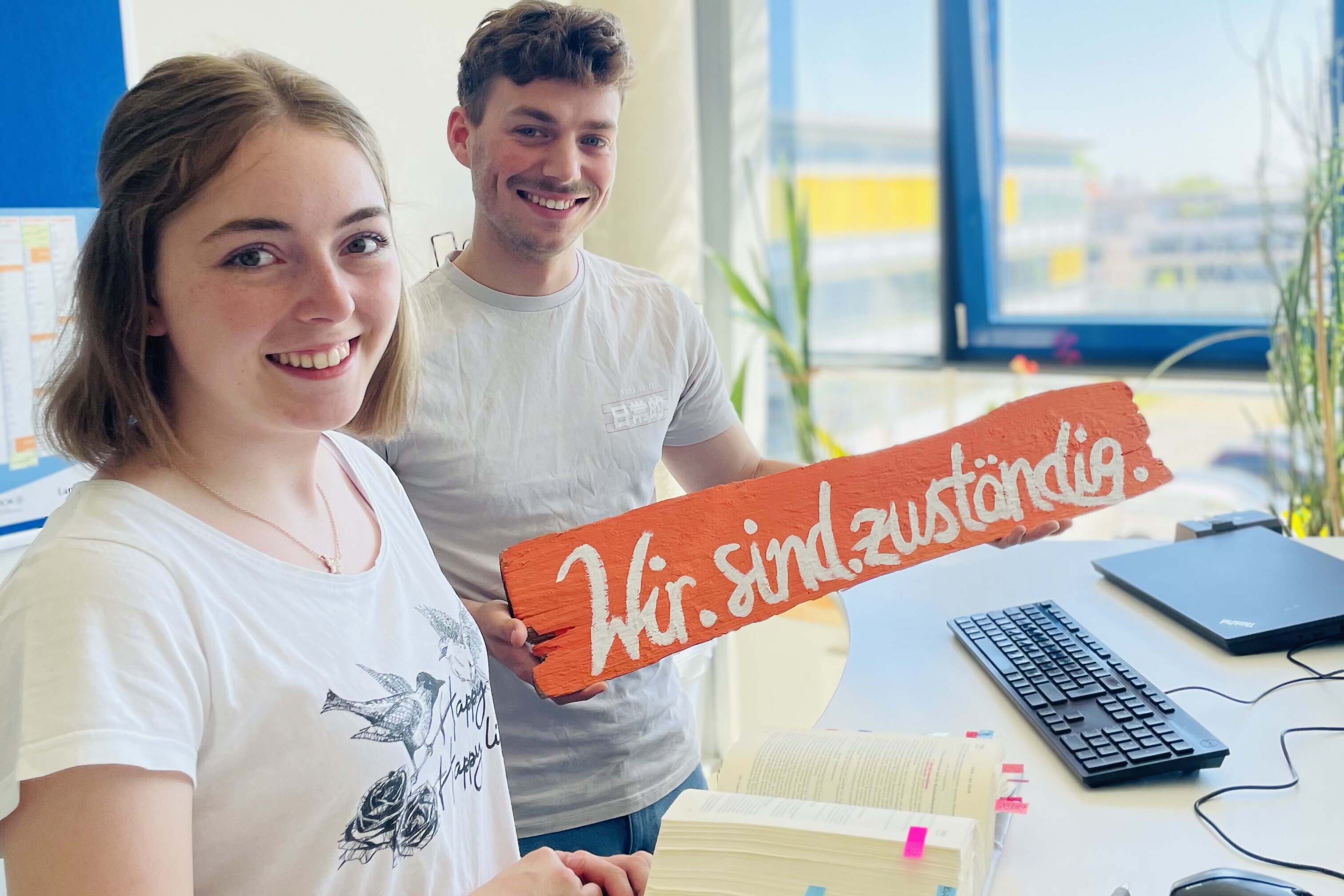 Zwei junge Menschen halten im Büro ein Schild, mit der Aufschrift "Wir sind zuständig", hoch.