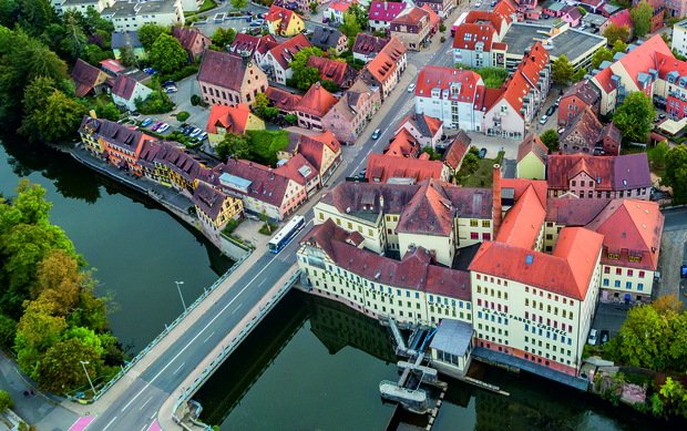 Luftbild der Stadt Stein mit einer idyllischen Ansicht der umliegenden Gebäude. In der Bildmitte das markante Gebäude der Fertigung und Akademie Faber-Castell, direkt am Fluss gelegen. Zu sehen ist der breite Flusslauf der Rednitz, der sich am Stadtrand erstreckt. Drumherum sind die Dächer der Wohnhäuser, grüne Flächen und Straßen zu sehen.