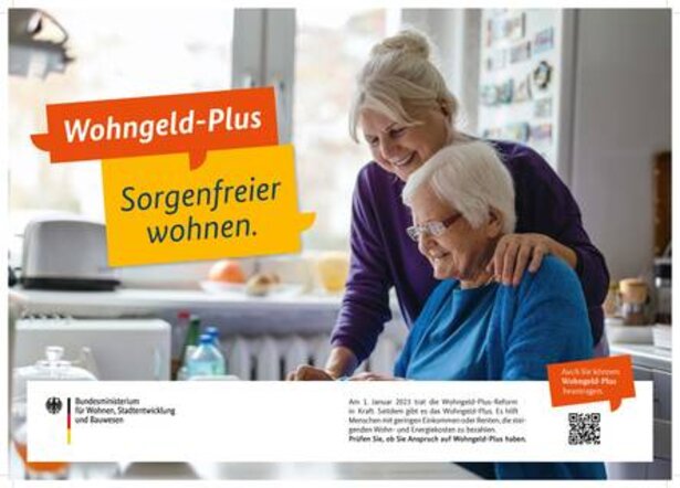 Eine ältere Frau sitzt am Tisch. Eine weitere Frau hat ihren Arm um ihre Schultern gelegt. Beide lächeln und wirken glücklich.