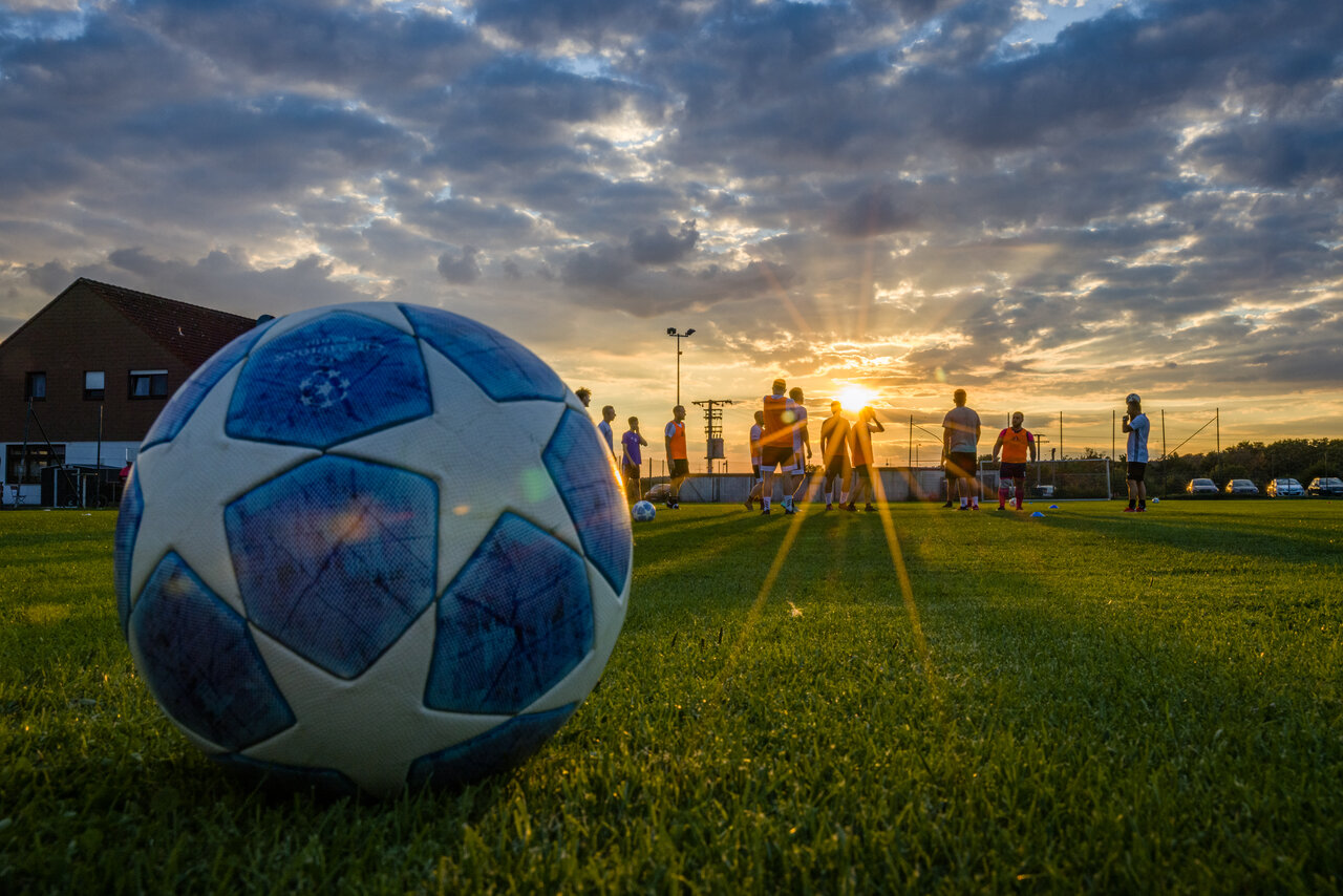 Das Bild zeigt einen Sportplatz mit einem Fußball, Jugendlichen im Hintergrund und einen Sonnenuntergang.