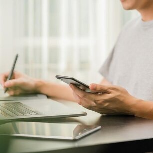 Eine Frau sitzt am Schreibtisch mit einem Handy in der linken Hand und einem Stift in der rechten Hand und schreibt etwas 