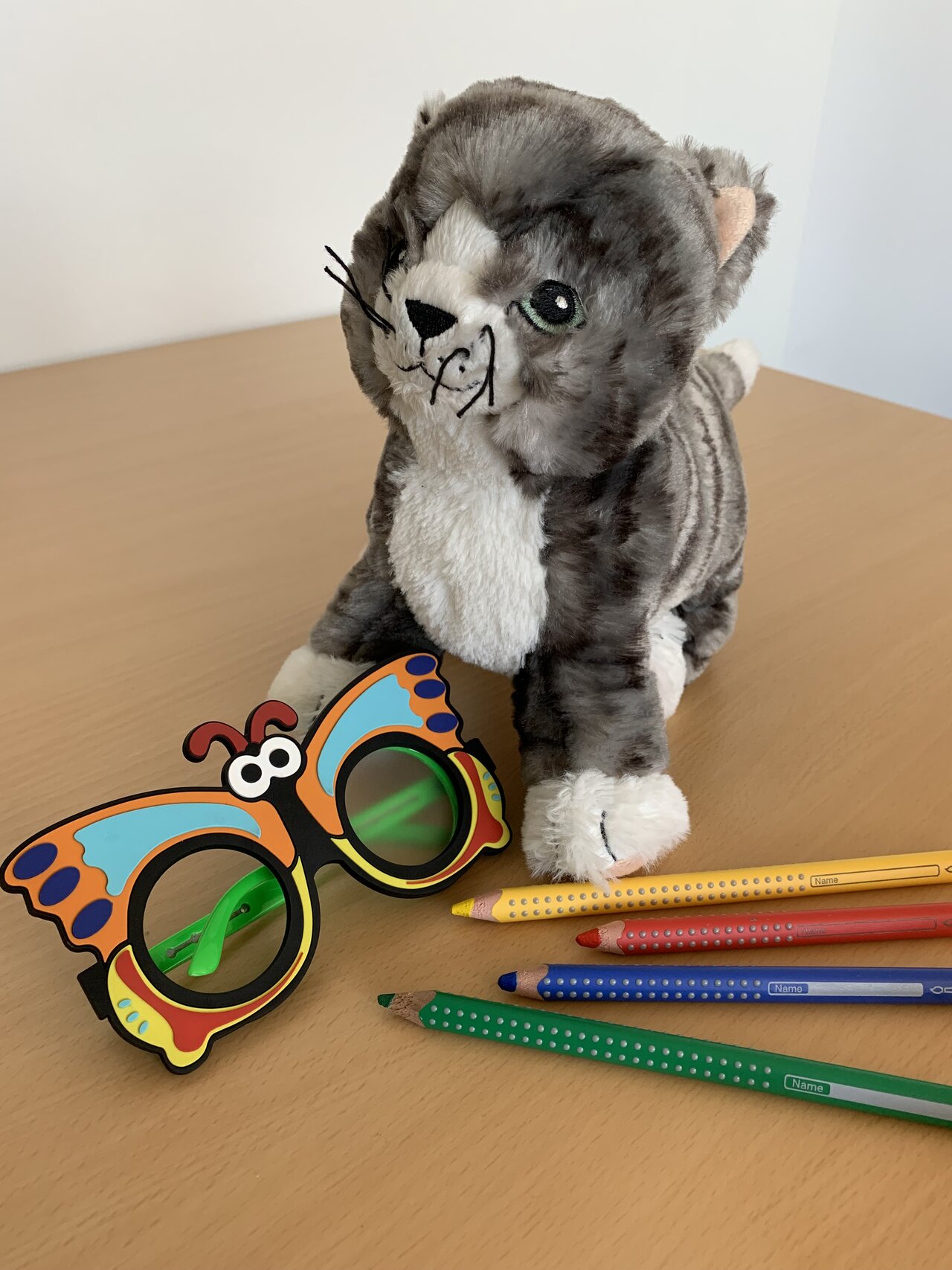 Verschiedene Utensilien der Schuleingangsuntersuchung - eine Plüschkatze, Buntstifte und eine Brille in Schmetterlingsform