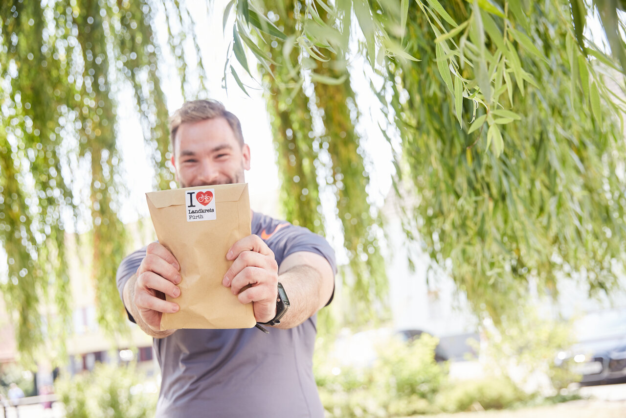 Ein lächelnder Mann steht im Freien unter einem Weidenbaum und hält eine braune Papiertüte mit beiden Händen in die Kamera. Auf der Tüte befindet sich ein Aufkleber mit dem Text "I love Landkreis Fürth" und einem roten Fahrradsymbol.