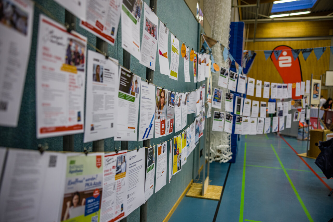 In einer Sporthalle sind beim "Tag der Ausbildung im Landkreis Fürth" zahlreiche Stellenbeschreibungen auf Papier ausgedruckt und an Leinen befestigt. Die Blätter hängen dicht nebeneinander und informieren über verschiedene Praktika- sowie Ausbildungsstellen. Im Hintergrund sind Messestände zu sehen. Die Veranstaltung bietet Jugendlichen einen Überblick über attraktive Karrierechancen in der Region.