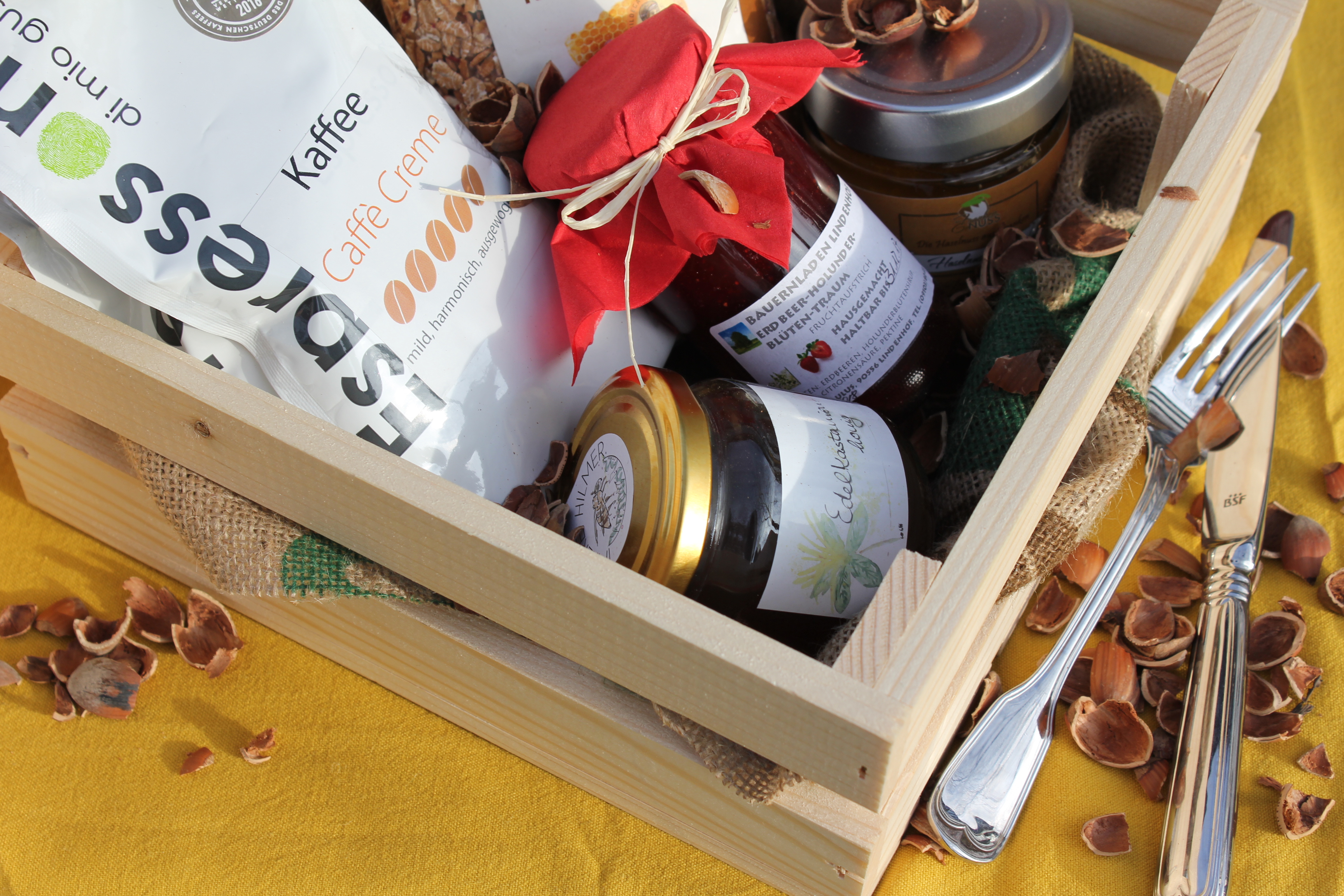 Eine Holzkiste, gefüllt mit verschiedenen regionalen oder handgemachten Produkten, steht auf einem gelben Tuch. In der Kiste befinden sich eine Packung Kaffee, mehrere Gläser mit Marmelade und Honig dekorativ verpackt. Um die Kiste herum liegen Besteckteile und verstreute Nussschalen.